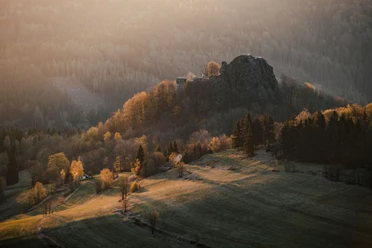 Burg Tollenstein Hügelige Landschaft bei Sonnenuntergang mit einer Burgruine auf einem bewaldeten Hügel, umgeben von herbstlichen Bäumen und sanften Lichtstrahlen.