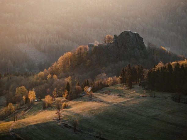 Burg Tollenstein Hügelige Landschaft bei Sonnenuntergang mit einer Burgruine auf einem bewaldeten Hügel, umgeben von herbstlichen Bäumen und sanften Lichtstrahlen.