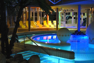 Beleuchtetes Schwimmbecken bei Nacht in einer modernen Spa-Anlage mit gelben Liegestühlen im Hintergrund.