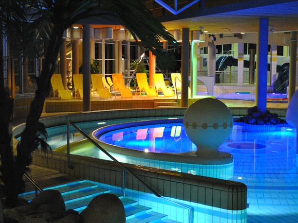 Beleuchtetes Schwimmbecken bei Nacht in einer modernen Spa-Anlage mit gelben Liegestühlen im Hintergrund.