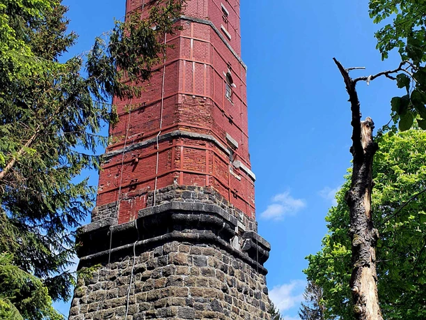 A tall, red-brick tower with a stone base stands surrounded by trees under a clear blue sky.