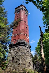 Tanzplan Ein hoher, roter Backsteinturm mit steinernem Sockel steht umgeben von Bäumen unter klarem, blauem Himmel.