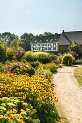 Hofcafé Abtsküche in Heiligenhaus Ein Weg führt durch einen üppig bepflanzten Garten zu einem Fachwerkhaus unter klarem Himmel.