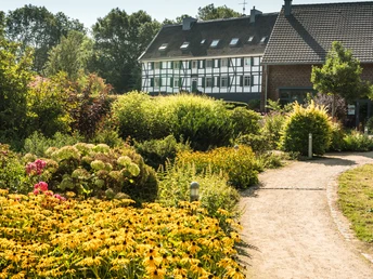 Hofcafé Abtsküche in Heiligenhaus Ein Weg führt durch einen üppig bepflanzten Garten zu einem Fachwerkhaus unter klarem Himmel.