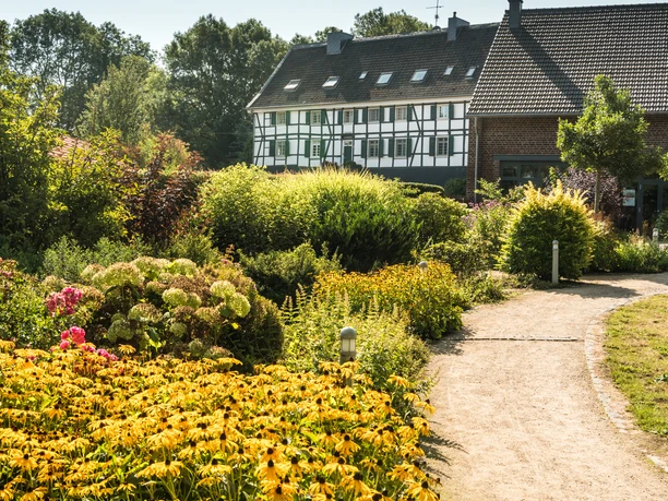 Hofcafé Abtsküche in Heiligenhaus Ein Weg führt durch einen üppig bepflanzten Garten zu einem Fachwerkhaus unter klarem Himmel.