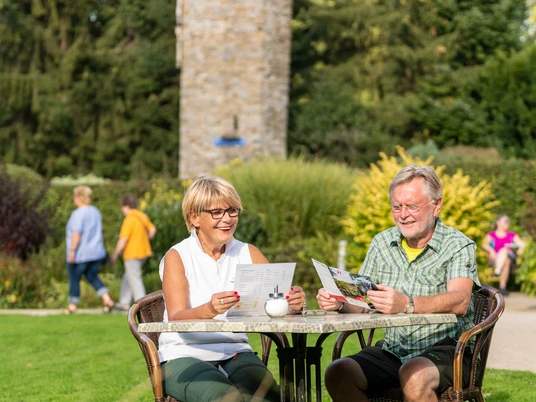 am Hofcafé Abtsküche in Heiligenhaus Ein älteres Paar liest entspannt an einem Tisch im grünen Garten vor einem steinernen Turm.
