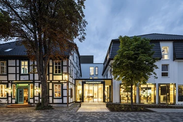 Hotelansicht Hotel WunnersWat Beleuchtetes modernes Glasgebäude mit traditionellen Fachwerkhäusern bei Dämmerung umgeben von Bäumen.