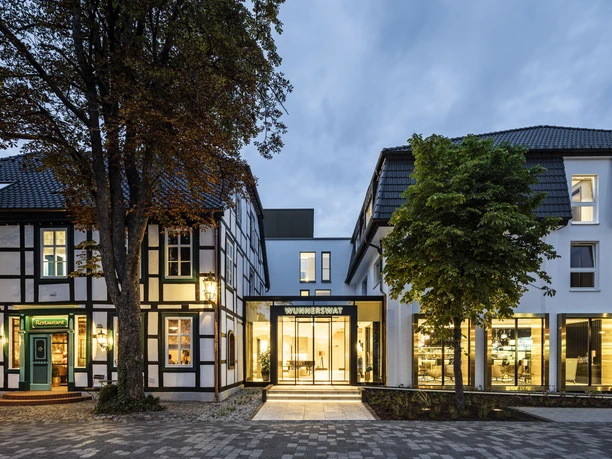 Hotelansicht Hotel WunnersWat Beleuchtetes modernes Glasgebäude mit traditionellen Fachwerkhäusern bei Dämmerung umgeben von Bäumen.