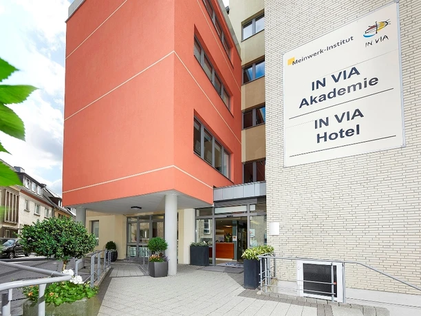 Moderne Hotelansicht mit orangefarbener Fassade und Eingangsbereich, umgeben von städtischer Umgebung.