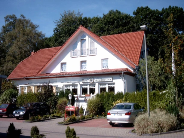 Weißes Gebäude mit rotem Giebeldach, umgeben von Pflanzen, mit Parkplatz und eingangsseitigen Fenstern.