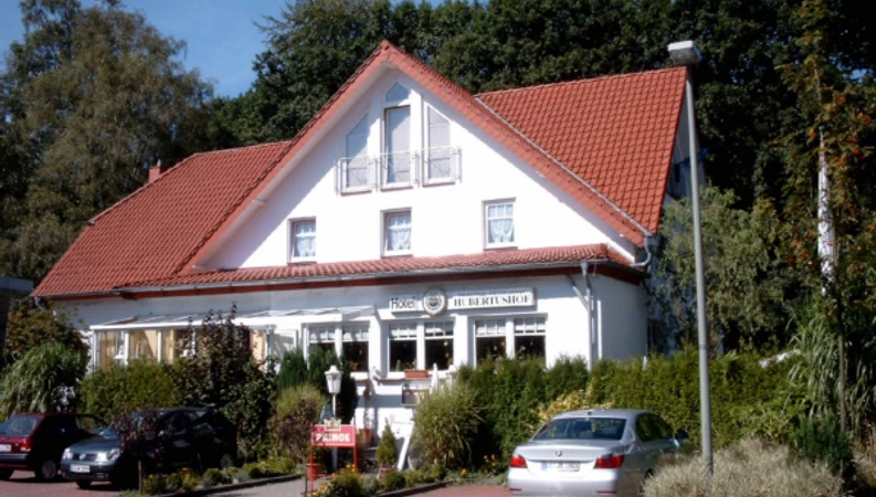 Hotel Restaurant Hubertushof Weißes Gebäude mit rotem Giebeldach, umgeben von Pflanzen, mit Parkplatz und eingangsseitigen Fenstern.