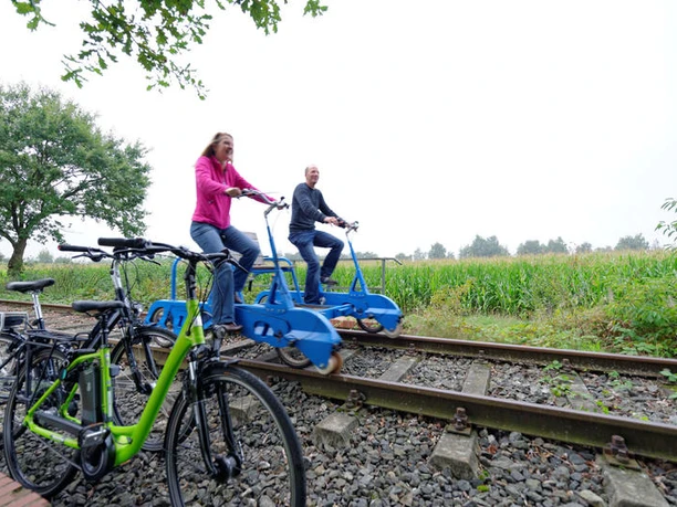 Zwei Personen fahren mit blauen Draisinen auf Bahngleisen durch eine grüne Landschaft neben Fahrrädern.