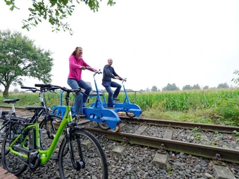 Draisinen Zwei Personen fahren mit blauen Draisinen auf Bahngleisen durch eine grüne Landschaft neben Fahrrädern.