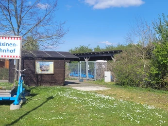 Blauer Draisinenschienenwagen auf Rasen vor einem kleinen Holzgebäude mit Draisinen-Bahnhof-Schild.