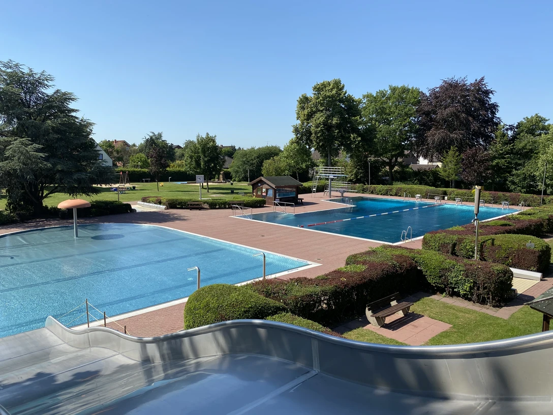 Ein Freibad mit zwei Schwimmbecken, umgeben von Rasenflächen und Bäumen an einem sonnigen Tag.