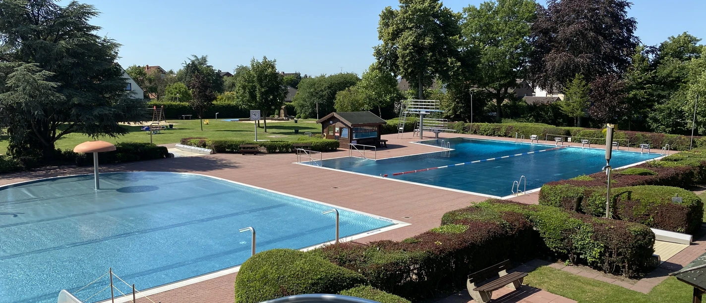 Ein Freibad mit zwei Schwimmbecken, umgeben von Rasenflächen und Bäumen an einem sonnigen Tag.