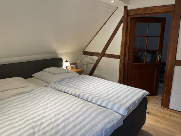 Gemütliches Schlafzimmer mit Doppelbett, Holzbalken an der Wand und warmem Licht, im Landhausstil.