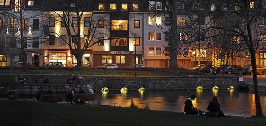 Abendstimmung am Flussufer vor dem beleuchteten Hotel Irma, Menschen sitzen entspannt auf Bänken.