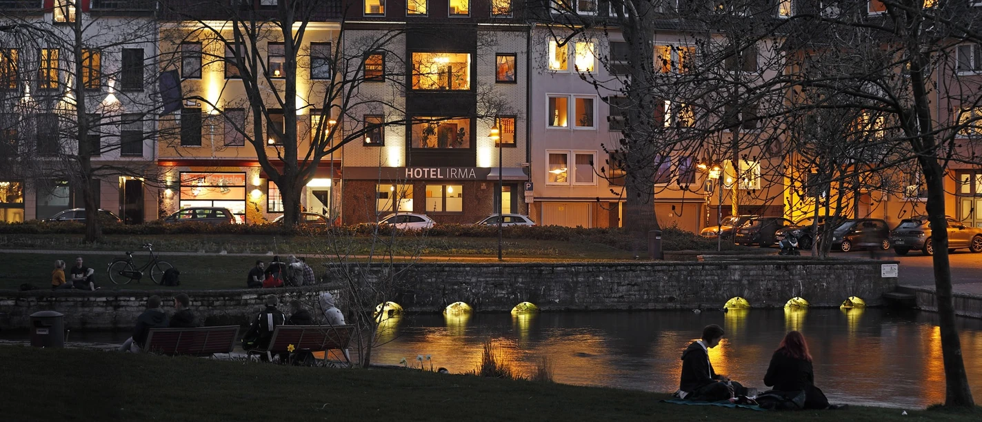 Abendstimmung am Flussufer vor dem beleuchteten Hotel Irma, Menschen sitzen entspannt auf Bänken.