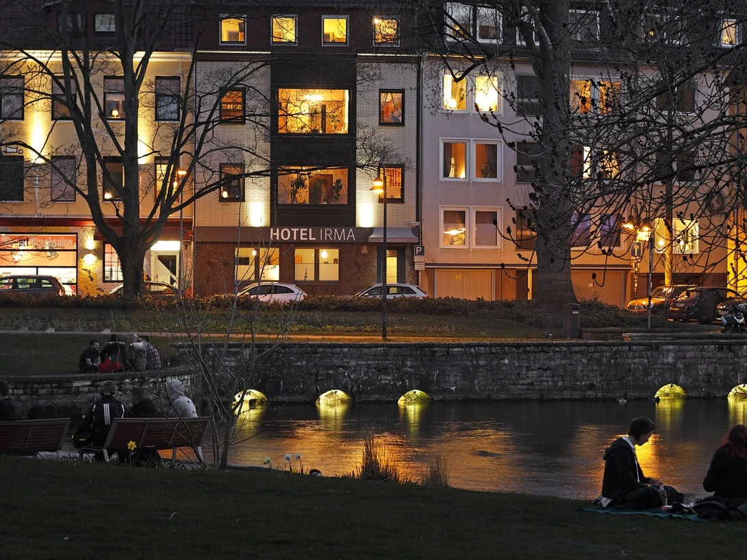 Hotel Irma Abendstimmung am Flussufer vor dem beleuchteten Hotel Irma, Menschen sitzen entspannt auf Bänken.