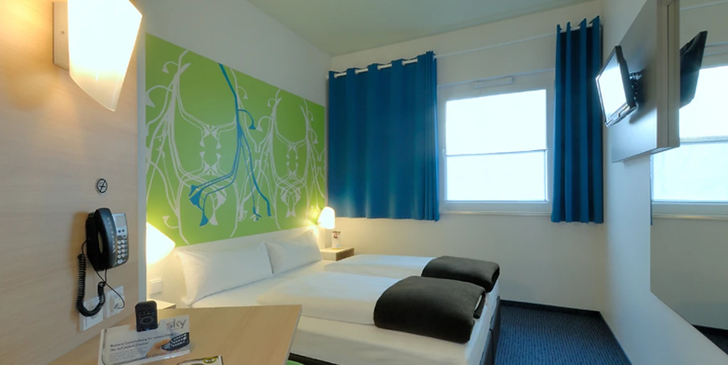 Zweibettzimmer mit modernen Möbeln, blauer Teppich, farbige Akzente und Flachbildfernseher.