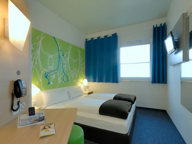 Zweibettzimmer mit modernen Möbeln, blauer Teppich, farbige Akzente und Flachbildfernseher.