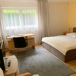 Helles, freundliches Gästezimmer mit Doppelbett, Schreibtisch und großem Fenster, Gardinen rahmen.