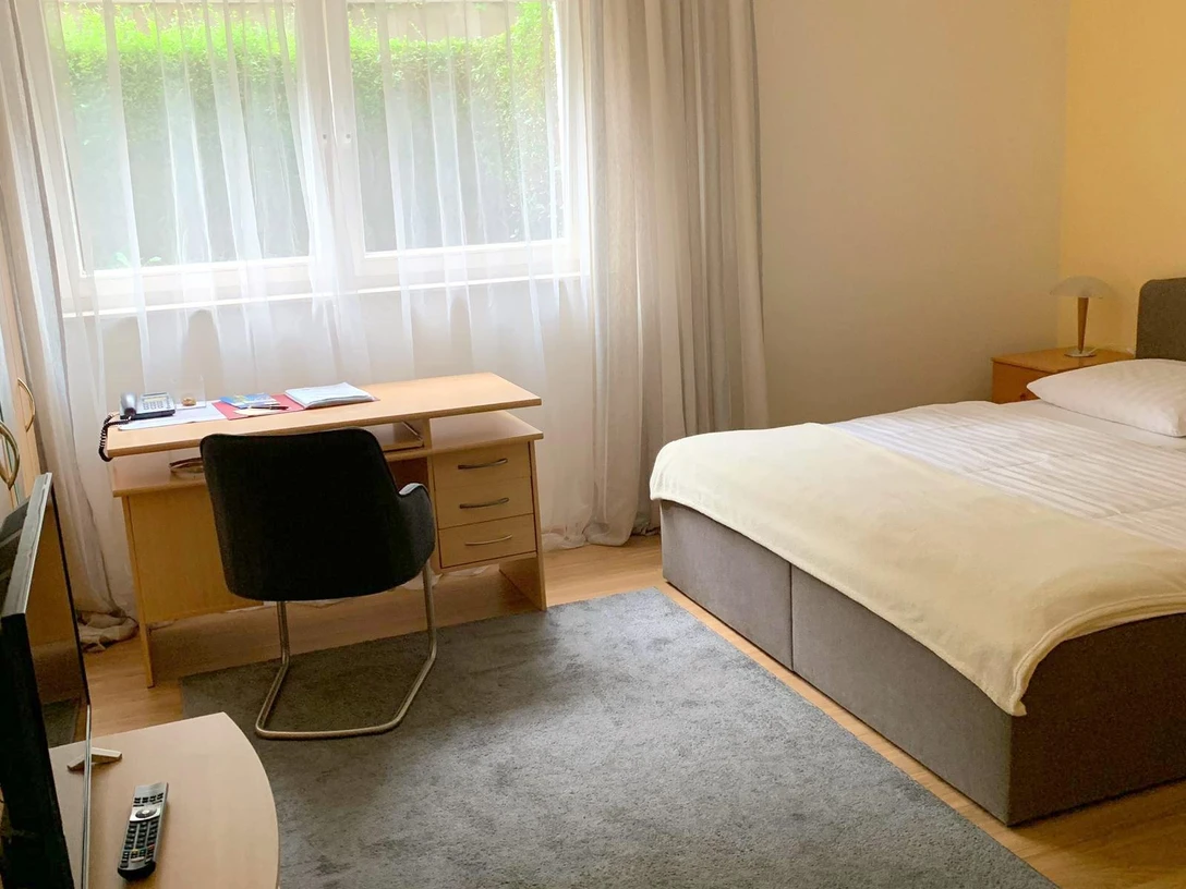Pension Kluth Helles, freundliches Gästezimmer mit Doppelbett, Schreibtisch und großem Fenster, Gardinen rahmen.