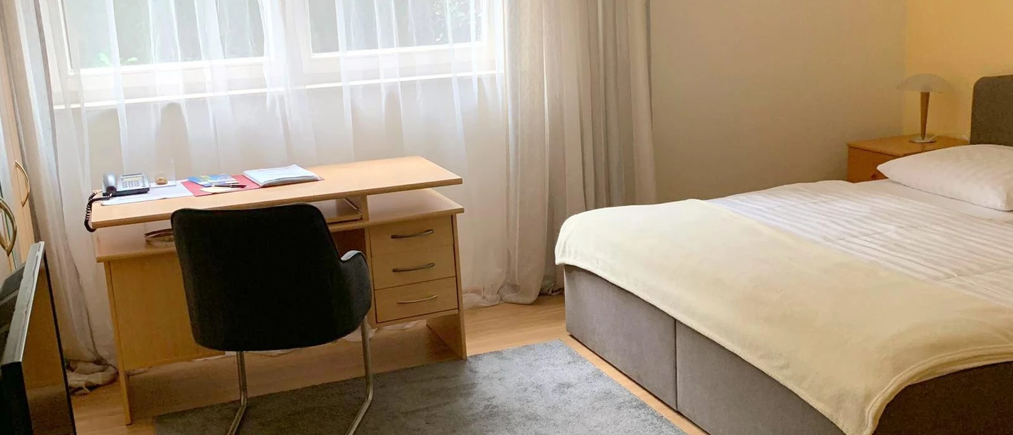 Pension Kluth Helles, freundliches Gästezimmer mit Doppelbett, Schreibtisch und großem Fenster, Gardinen rahmen.