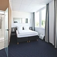 Helles Hotelzimmer mit Doppelbett, Teppichboden, großen Fenstern und angrenzendem Bad mit Glaswand.