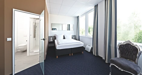 Road House Hotel Helles Hotelzimmer mit Doppelbett, Teppichboden, großen Fenstern und angrenzendem Bad mit Glaswand.