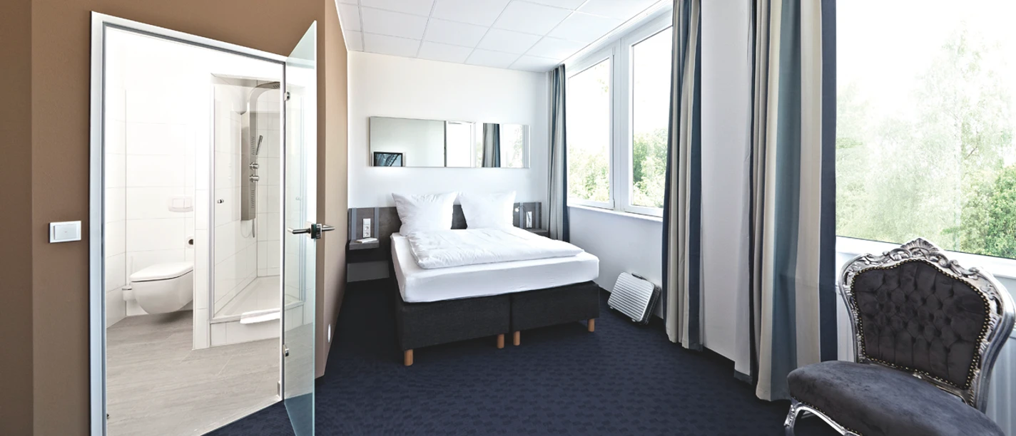 Road House Hotel Helles Hotelzimmer mit Doppelbett, Teppichboden, großen Fenstern und angrenzendem Bad mit Glaswand.