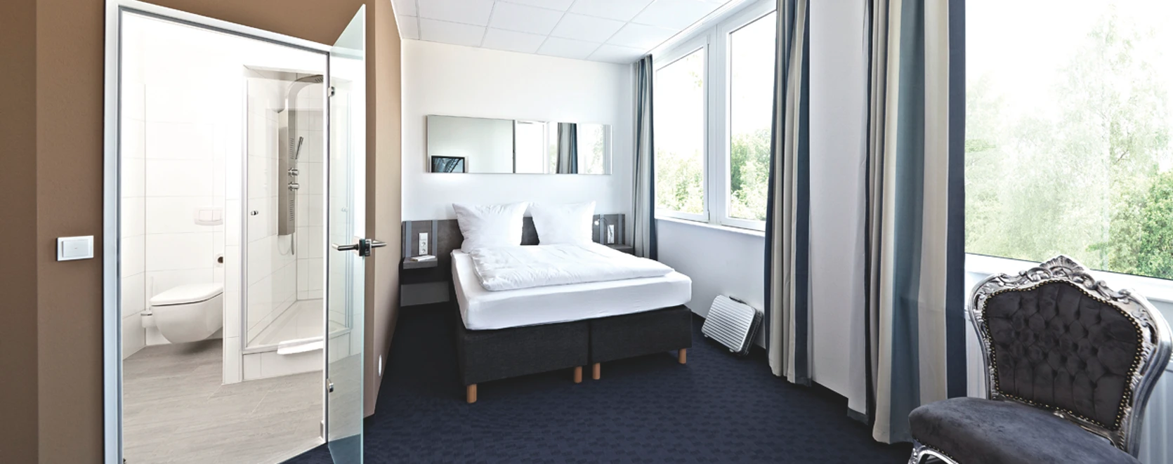 Road House Hotel Helles Hotelzimmer mit Doppelbett, Teppichboden, großen Fenstern und angrenzendem Bad mit Glaswand.