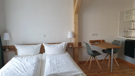 Das grüne Haus Helles Zimmer mit Doppelbett, Holzbalken, Esstisch, zwei Stühlen und schlichter Küchenecke.