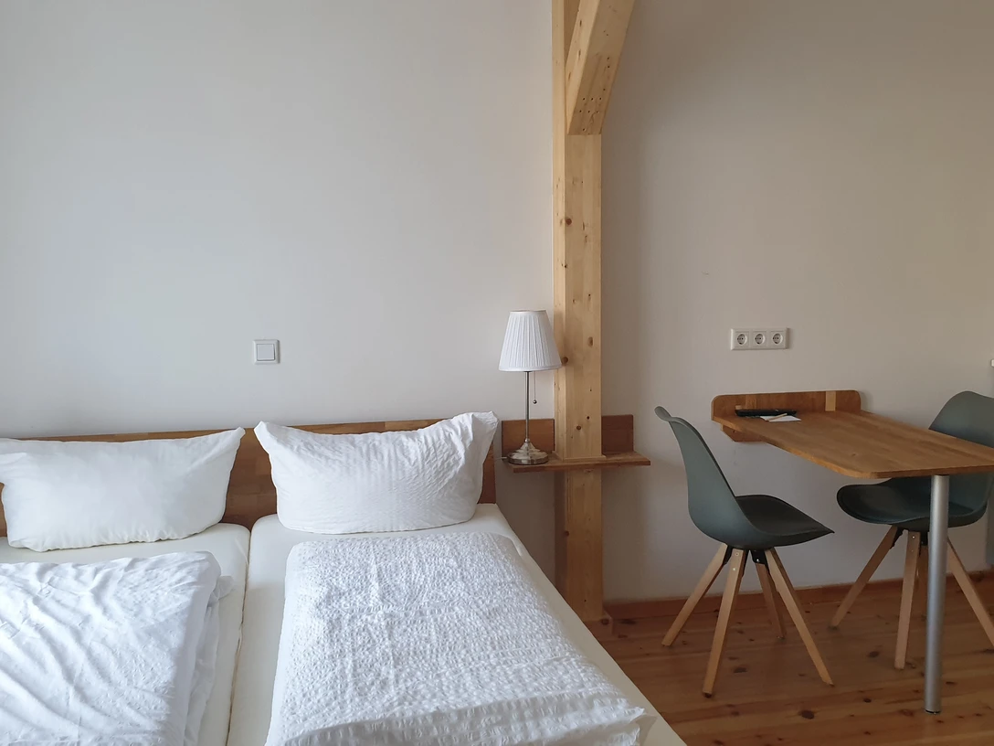Das grüne Haus Helles Zimmer mit Doppelbett, Holzbalken, Esstisch, zwei Stühlen und schlichter Küchenecke.