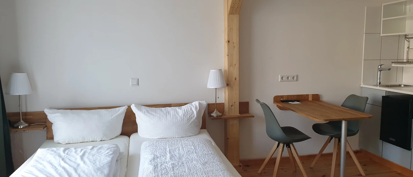 Das grüne Haus Helles Zimmer mit Doppelbett, Holzbalken, Esstisch, zwei Stühlen und schlichter Küchenecke.