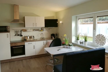 Moderne Küche in einer Ferienwohnung mit Esstisch, weißen Schränken und Blick auf ein Fenster.