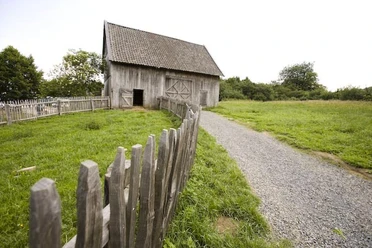 Gartenmarkt im Freilichtmuseum Ein alter Holzschuppen steht auf einer grünen Wiese mit einem Weg und Holzzaun im Vordergrund.