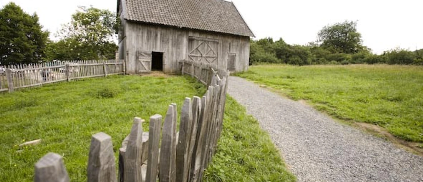 Gartenmarkt im Freilichtmuseum Ein alter Holzschuppen steht auf einer grünen Wiese mit einem Weg und Holzzaun im Vordergrund.