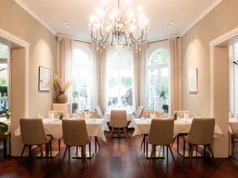 Villa Q Innenräume Die helle Inneneinrichtung der Villa Q entfaltet sich in einem harmonischen Zusammenspiel aus warmem Sonnenlicht, das sanft in den offenen Raum strömt. Die eleganten Tische und Stühle in zarten Beige- und Weißtönen schaffen eine angenehme Atmosphäre, während ein prächtiger Kronleuchter von der Decke herabstrahlt und dem Raum einen Hauch von Luxus verleiht.