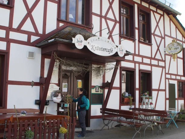 Kräutercafé <p>Ein Fachwerkhaus mit dem Schild "Kräutercafé", geschmückt mit Pflanzen, steht an einer Straße.</p>