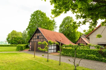 Alte Scheune Fachwerk-Scheune mit rotem Ziegeldach, umgeben von grüner Hecke und Bäumen, blauer Himmel.