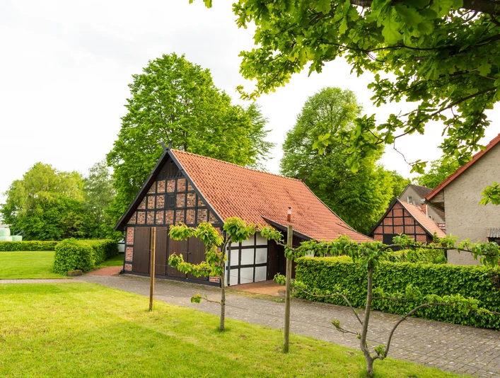 Alte Scheune Fachwerk-Scheune mit rotem Ziegeldach, umgeben von grüner Hecke und Bäumen, blauer Himmel.