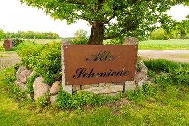 Holzhausen II Schild mit der Aufschrift "Alte Schmiede" vor einer grünen Landschaft mit Baum und Feld im Hintergrund.