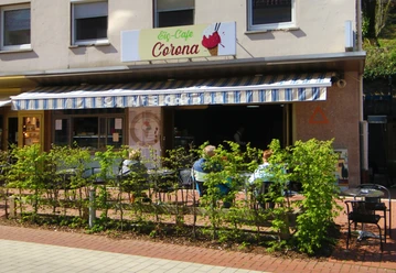 Eiscafé Corona Der Außenbereich des Eiscafés Corona. Man kann Stühle und Tische unter einer Markise durch eine Hecke hindurch sehen
