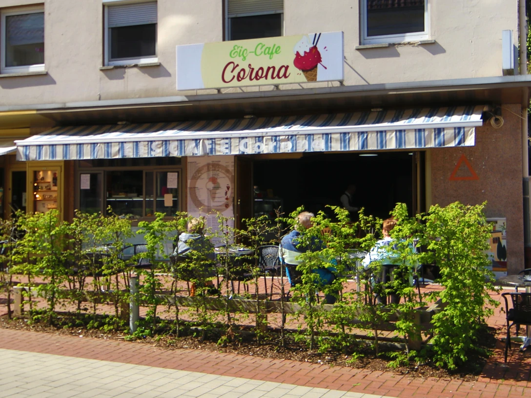 Eiscafé Corona Der Außenbereich des Eiscafés Corona. Man kann Stühle und Tische unter einer Markise durch eine Hecke hindurch sehen