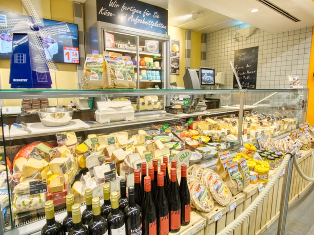Eine einladende Käsetheke zeigt verschiedene Käsesorten, Flaschen Rotwein und Backwaren.