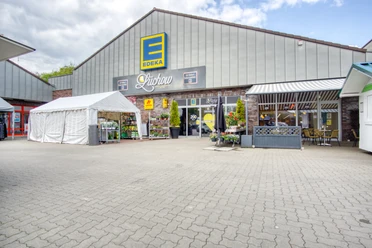 Außenansicht Edeka Lüchow Die Außenansicht des Edeka-Marktes in Lüchow zeigt einen weitläufigen Parkplatz vor einem modernen Ladengebäude. Das flache Dach und die großen Glasfenster prägen das Erscheinungsbild.