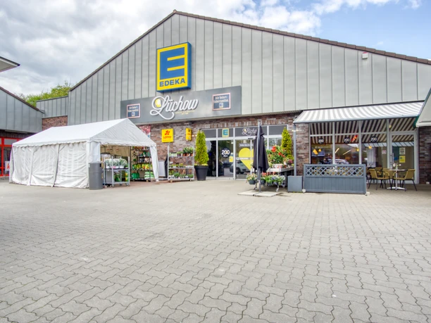 Außenansicht Edeka Lüchow Die Außenansicht des Edeka-Marktes in Lüchow zeigt einen weitläufigen Parkplatz vor einem modernen Ladengebäude. Das flache Dach und die großen Glasfenster prägen das Erscheinungsbild.