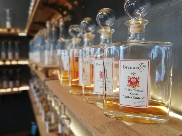 BrennerEy Flaschen mit verschiedenen Spirituosen auf Holzregalen, beschriftet mit "BrennerEy" und Geschmacksrichtungen.